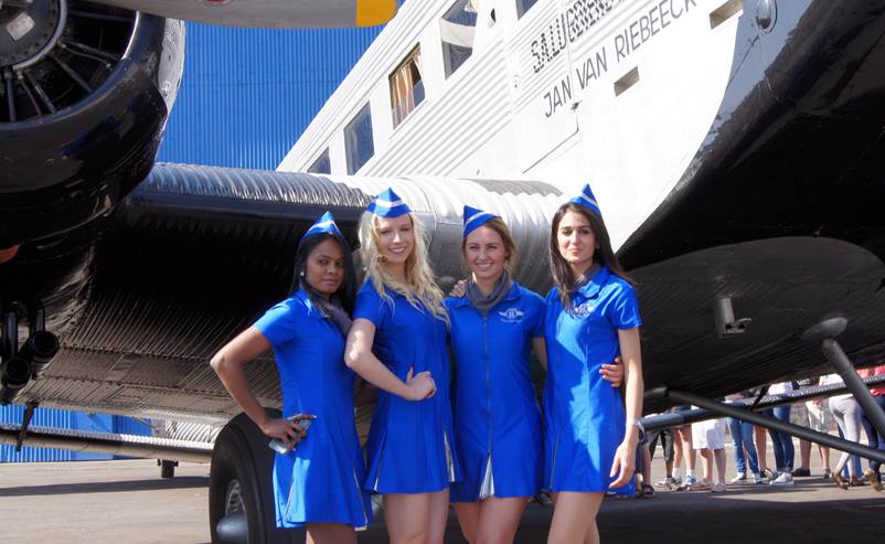 A group of women in blue dresses and hats standing in front of an airplane

Description automatically generated